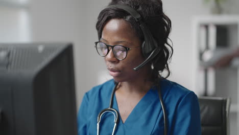 African-american-black-female-general-practitioner-in-white-coat-sitting-at-desk-in-doctor's-office-and-scrolling-computer-mouse-while-reading-patient's-medical-history